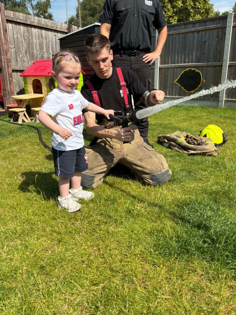 Child at Crow Kids Day Nursery in Walsall in the nursery garden holds a fire fighter hose with water shooting out, next to a fire fighter helping her to point the hose in the correct direction